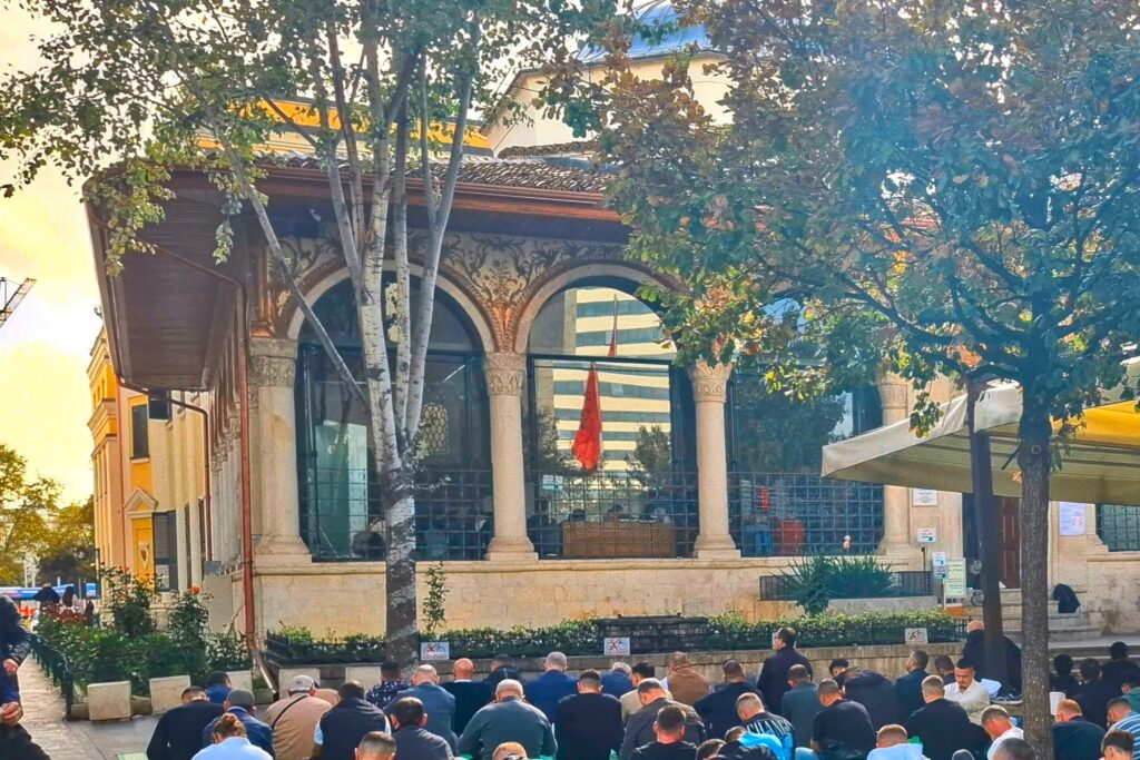 מסגד האטם ביי עם מתפללים יושבים בחוץ ובפנים דגל אלבניה פיתוחי הציורים כיכר סקנדרבג